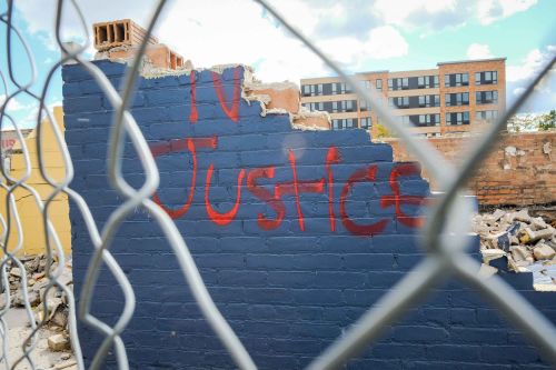 part of a demolished wall stands in view through a fence with the word "justice" spray painted on it